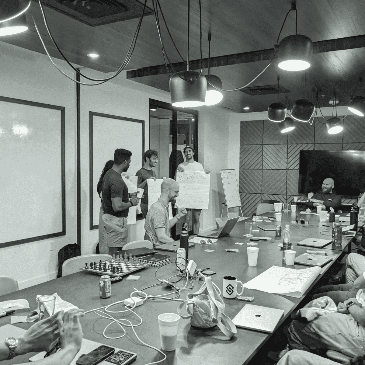 Group of people in a meeting room with whiteboards, laptops, and a chessboard on a table, discussing ideas while holding presentation boards.